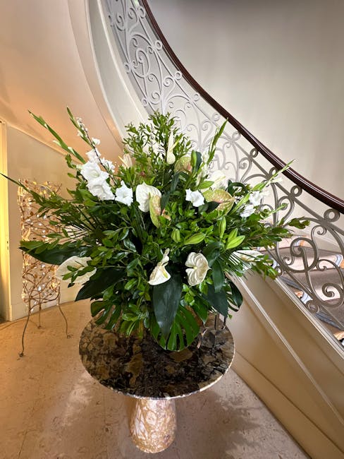 A large, elegant floral arrangement featuring white calla lilies, anthuriums, and various green foliage, arranged in a black vase. The bouquet is placed on a round, dark marble table near a curved sta