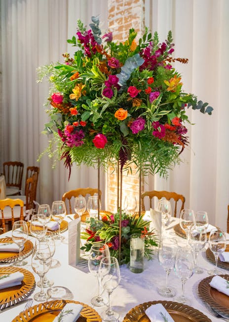 A large, vibrant floral arrangement featuring a mix of colorful flowers such as pink and red roses, purple snapdragons, yellow and orange blooms, and lush green foliage, displayed on a tall, slender s