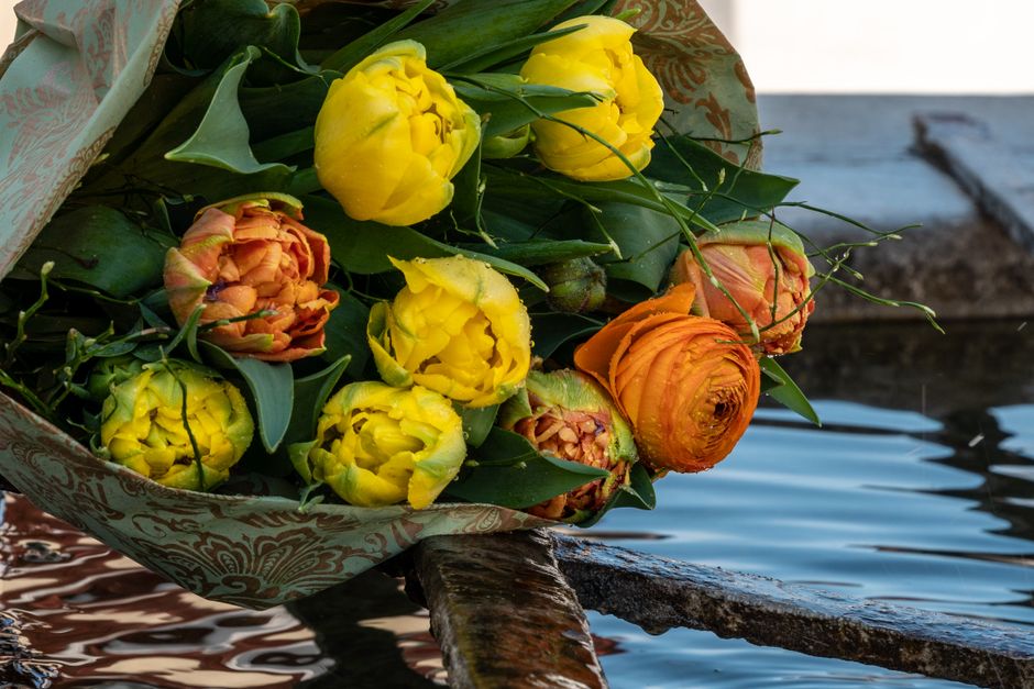 A bouquet of fresh yellow and orange ranunculus flowers with lush green leaves, wrapped in decorative patterned paper, is resting on a weathered wooden dock near a calm body of water. The vivid blooms