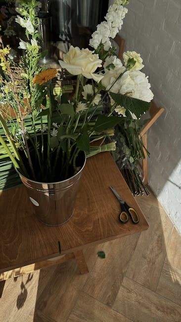 A tall floral arrangement placed in a silver bucket on a wooden table, featuring large white roses, white stock flowers, and smaller yellow and white blooms with lush green foliage. The bouquet appear
