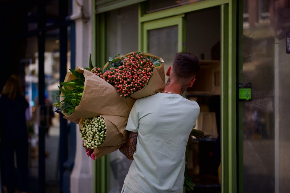 A person with short dark hair, wearing a light-colored T-shirt, is holding a large bouquet of fresh flowers wrapped in brown paper with green foliage peeking out. The bouquet includes clusters of pink