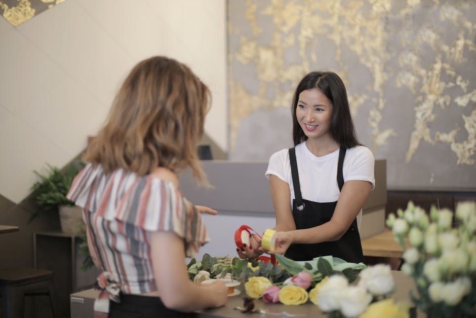 A cheerful female florist with long dark hair, wearing a white t-shirt and black apron, is smiling as she hands over a floral gift bouquet to a customer at a flower shop. The bouquet includes vibrant 