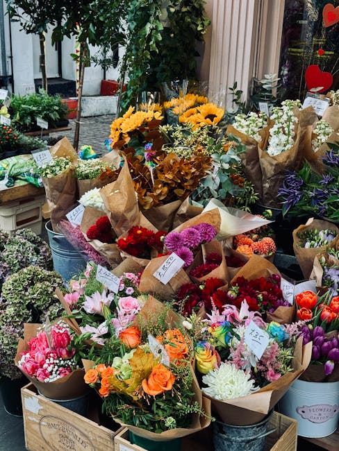 A vibrant display of freshly cut flowers arranged in bouquets and potted plants at a flower shop's outdoor stand, including bright yellow sunflowers, pink lilies, red roses, purple tulips, and white c