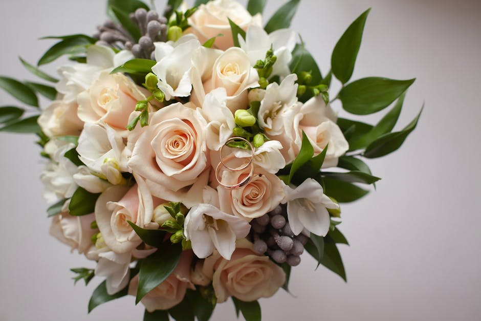 A close-up view of a floral bouquet featuring pale blush roses, white freesias, and small green buds, arranged with dark green, elongated leaves. The flowers are fresh, with delicate, soft petals and