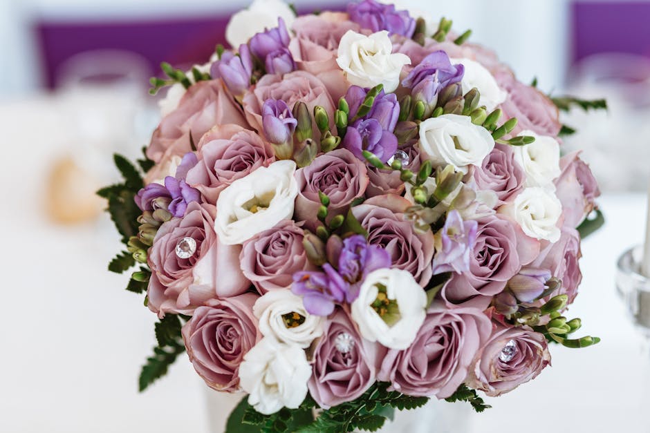 A close-up image of a floral bouquet featuring blush pink roses, white lisianthus, and purple freesia, arranged in a rounded style with visible green ferns and foliage at the base. The flowers appear